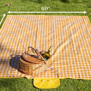 Puede incluir: Una manta de picnic a cuadros amarillos y blancos, de 152 x 198 cm, est&aacute; extendida sobre hierba verde. Una cesta de picnic de mimbre con una botella de vino y otros art&iacute;culos est&aacute; sobre la manta. Una peque&ntilde;a bolsa amarilla est&aacute; adjunta.