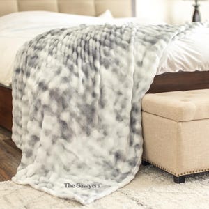 May include: A plush, oversized blanket in shades of gray and white drapes over a bed. The blanket has a soft, textured appearance. The Sawyer's logo is visible in the corner. A beige ottoman sits at the foot of the bed.