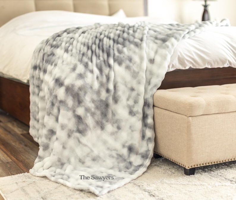 May include: A plush, oversized blanket in shades of gray and white drapes over a bed. The blanket has a soft, textured appearance. The Sawyer's logo is visible in the corner. A beige ottoman sits at the foot of the bed.