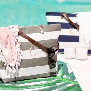May include: Two striped canvas tote bags with leather handles. The bag on the left is gray and white with a pink scarf and sunglasses inside. The bag on the right is navy and white with a white scarf inside. Both bags have a monogrammed initial on the front.