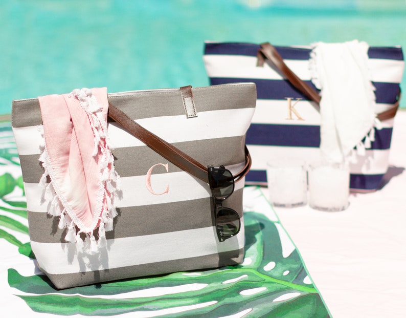 May include: Two striped canvas tote bags with leather handles. The bag on the left is gray and white with a pink scarf and sunglasses inside. The bag on the right is navy and white with a white scarf inside. Both bags have a monogrammed initial on the front.