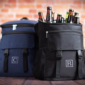 K&ouml;nnte beinhalten: Zwei schwarze und blaue Rucks&auml;cke mit isolierten F&auml;chern. Der schwarze Rucksack hat eine Rei&szlig;verschlusstasche und ist mit braunen Glasflaschen gef&uuml;llt. Der blaue Rucksack hat einen wei&szlig;en Buchstaben "C" auf der Vorderseite gestickt. Der schwarze Rucksack hat einen wei&szlig;en Buchstaben "R" auf der Vorderseite gestickt.