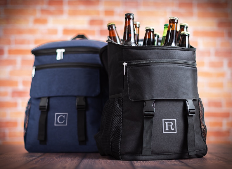 K&ouml;nnte beinhalten: Zwei schwarze und blaue Rucks&auml;cke mit isolierten F&auml;chern. Der schwarze Rucksack hat eine Rei&szlig;verschlusstasche und ist mit braunen Glasflaschen gef&uuml;llt. Der blaue Rucksack hat einen wei&szlig;en Buchstaben "C" auf der Vorderseite gestickt. Der schwarze Rucksack hat einen wei&szlig;en Buchstaben "R" auf der Vorderseite gestickt.