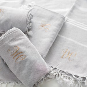 Puede incluir: Cuatro toallas grises con flecos blancos, cada una bordada con un conjunto diferente de iniciales en hilo dorado. Las iniciales son "BM", "DC", "LL" y "MC".