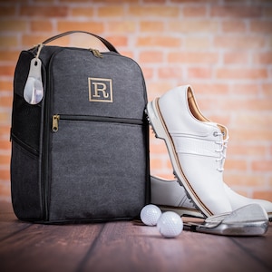 May include: A grey canvas shoe bag with a zipper and a leather handle. The bag has a monogrammed "R" on the front. Two white golf balls and a golf club are in the foreground.