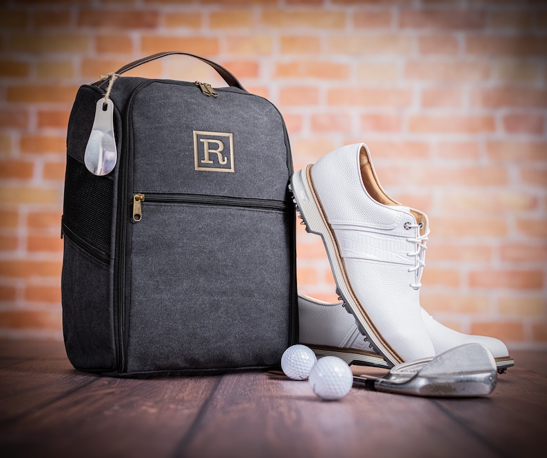 May include: A grey canvas shoe bag with a zipper and a leather handle. The bag has a monogrammed "R" on the front. Two white golf balls and a golf club are in the foreground.