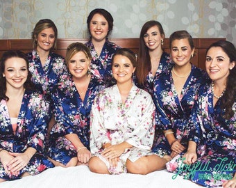 May include: Seven women wearing blue and white floral print robes. The robes have long sleeves and are tied at the waist. The women are sitting on a bed with a white bedspread.