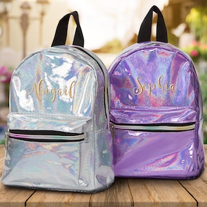May include: Two iridescent backpacks with personalized names. The backpack on the left is silver with the name Abigail embroidered in gold. The backpack on the right is purple with the name Sophia embroidered in gold.