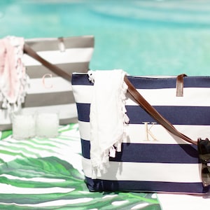 May include: Two striped canvas tote bags with leather handles. One bag is gray and white with the letter "C" embroidered on it. The other bag is navy blue and white with the letter "K" embroidered on it. Both bags have a white towel inside.