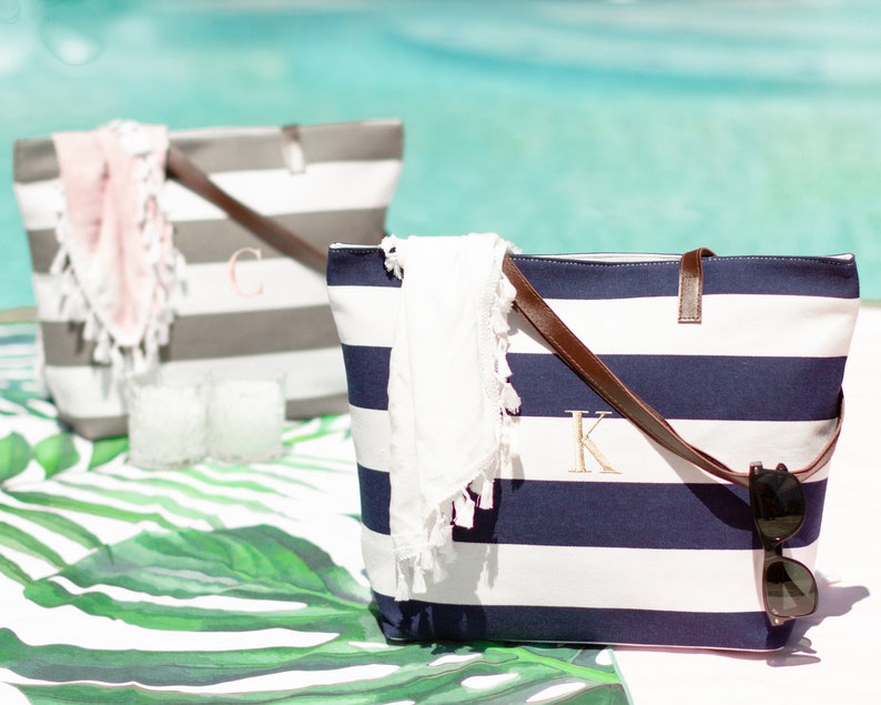 May include: Two striped canvas tote bags with leather handles. One bag is gray and white with the letter "C" embroidered on it. The other bag is navy blue and white with the letter "K" embroidered on it. Both bags have a white towel inside.