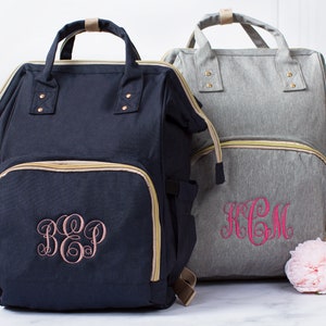 May include: Two backpacks with top handles and front zippered pockets. The navy blue backpack features a light pink embroidered monogram, while the heather gray backpack features a bright pink embroidered monogram. Both bags have gold-toned zippers.