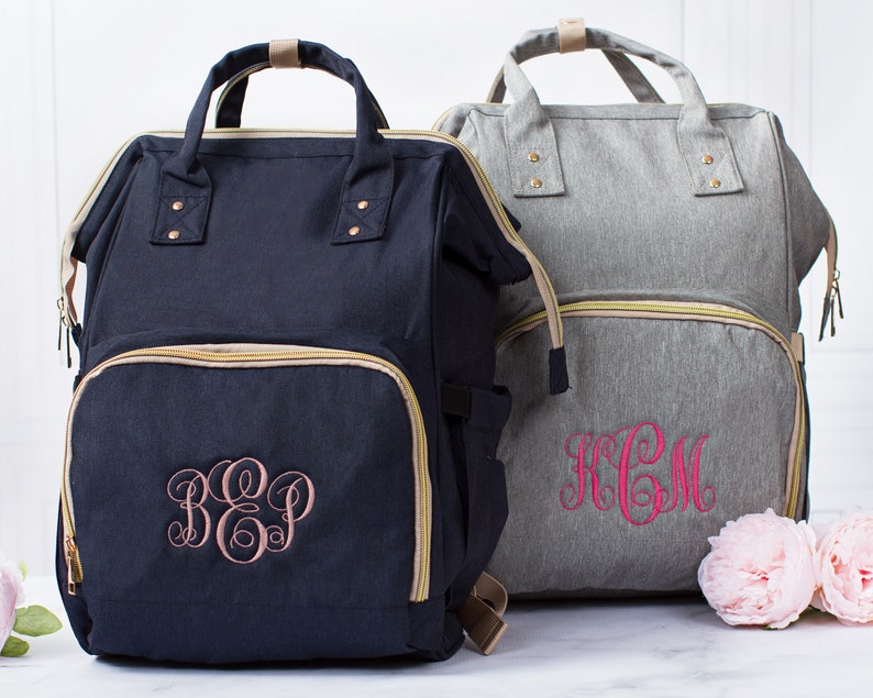 May include: Two backpacks with top handles and front zippered pockets. The navy blue backpack features a light pink embroidered monogram, while the heather gray backpack features a bright pink embroidered monogram. Both bags have gold-toned zippers.
