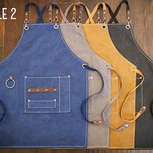 May include: Four canvas aprons in different colours: blue, grey, brown, and black. Each apron has adjustable straps, a large pocket, and a small loop for hanging tools. The aprons are labelled "STYLE 2".