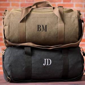 May include: Two canvas duffel bags, one tan and one black, stacked on top of each other. The tan bag has the embroidered initials "BM" and the black bag has "JD". Both bags have handles and shoulder straps.