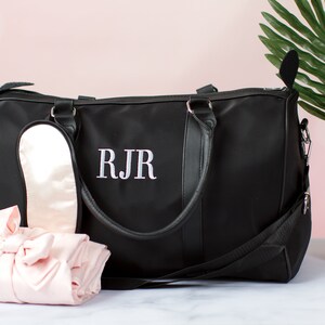 以下が含まれることがあります： 黒い旅行バッグは、黒い革のハンドルと白い刺繍のモノグラム「RJR」が付いています。バッグの隣には、ピンクのサテン製のアイマスクとピンクのサテン製のローブがあります。