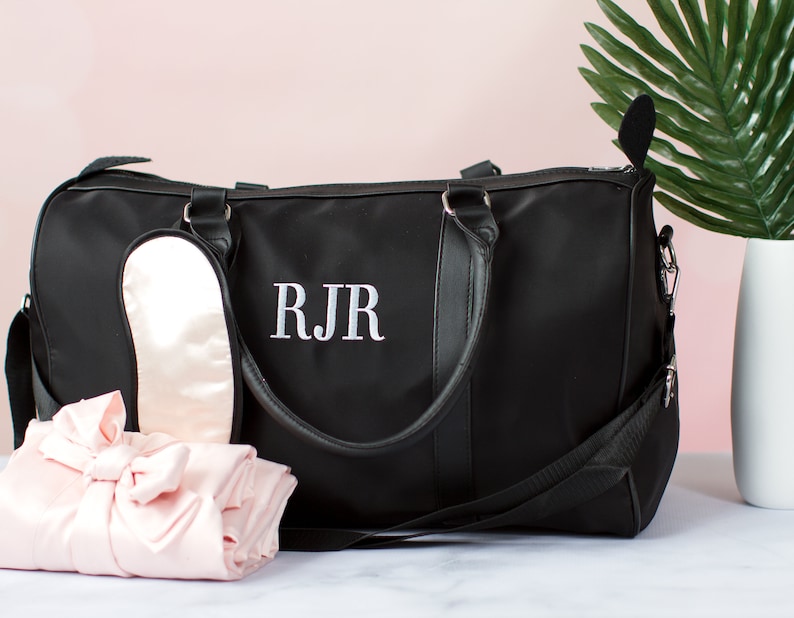 以下が含まれることがあります： 黒い旅行バッグは、黒い革のハンドルと白い刺繍のモノグラム「RJR」が付いています。バッグの隣には、ピンクのサテン製のアイマスクとピンクのサテン製のローブがあります。