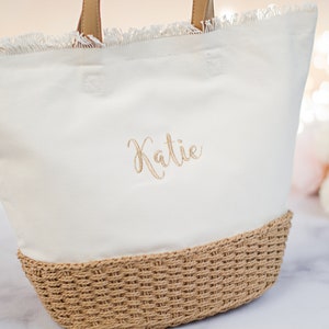 May include: White canvas tote bag with a frayed edge and a woven straw bottom. The bag has the name "Katie" embroidered on the front in gold thread.