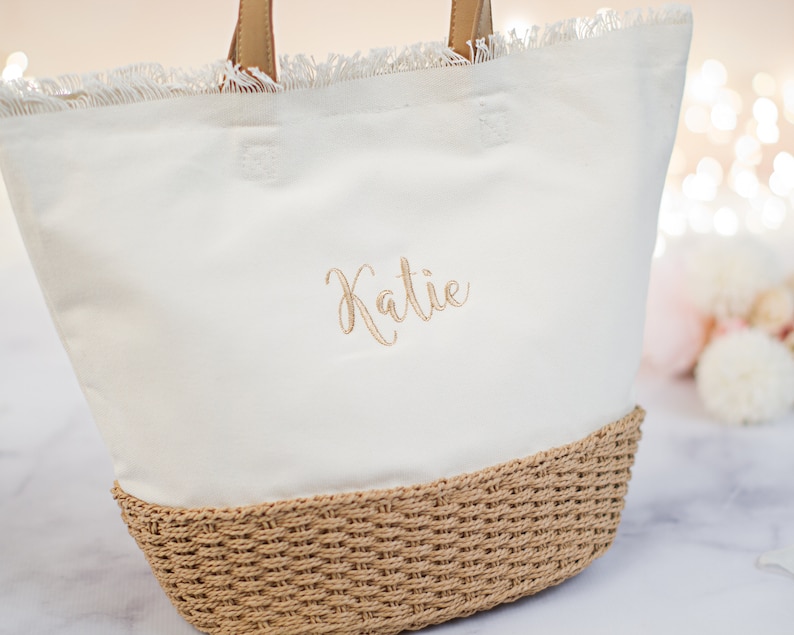 May include: White canvas tote bag with a frayed edge and a woven straw bottom. The bag has the name "Katie" embroidered on the front in gold thread.