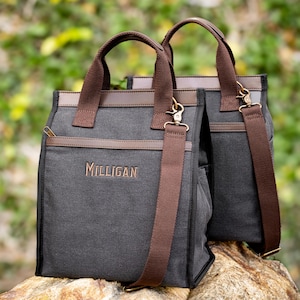 May include: Two charcoal gray tote bags with brown leather accents and straps. The bags have a rectangular shape, with a zipper closure and the name "Milligan" embroidered in gold. The bags have both handles and adjustable shoulder straps.