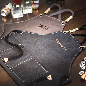May include: Two personalized black canvas aprons with adjustable straps and a large pocket. The aprons are embroidered with the names "DOYLE" and "FARRELL".
