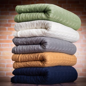 Pode incluir: Uma pilha de cinco cobertores de malha dobrados em v&aacute;rias cores, incluindo verde, branco, cinza, marrom e azul. Os cobertores est&atilde;o dispostos em uma pilha, com o cobertor verde no topo e o cobertor azul na parte inferior.
