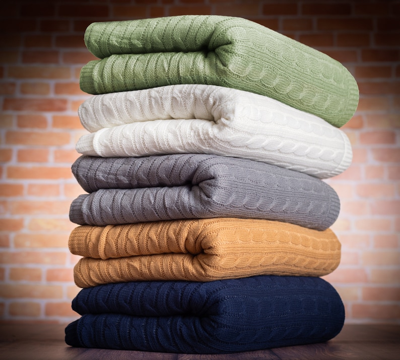 Pode incluir: Uma pilha de cinco cobertores de malha dobrados em v&aacute;rias cores, incluindo verde, branco, cinza, marrom e azul. Os cobertores est&atilde;o dispostos em uma pilha, com o cobertor verde no topo e o cobertor azul na parte inferior.