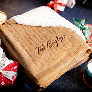 May include: A folded tan cable-knit blanket with a white sherpa lining. The blanket is embroidered with the name "The Bayley's" in a cursive font. The image includes lit candles and wrapped gifts, suggesting a cozy, gift-giving theme.