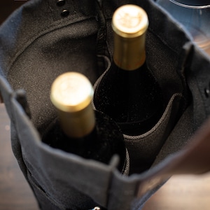 May include: Two wine bottles with gold tops are inside a black canvas tote bag with leather handles.