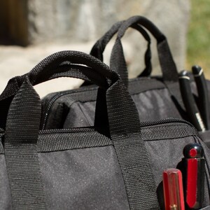 Puede incluir: Dos bolsas de herramientas de lona negra con m&uacute;ltiples bolsillos y cierres de cremallera. Una bolsa tiene una herramienta roja y negra en el bolsillo.