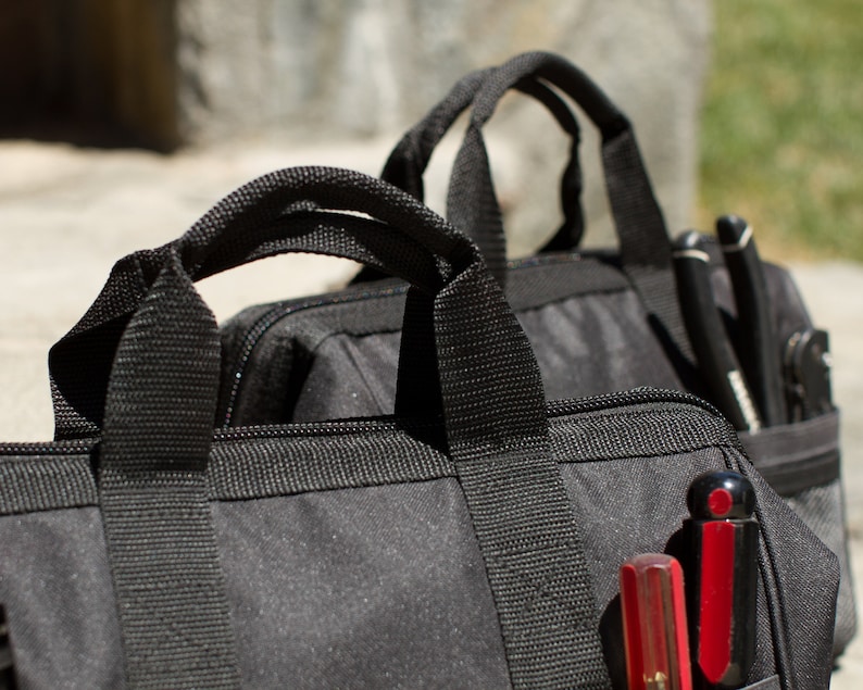 Puede incluir: Dos bolsas de herramientas de lona negra con m&uacute;ltiples bolsillos y cierres de cremallera. Una bolsa tiene una herramienta roja y negra en el bolsillo.