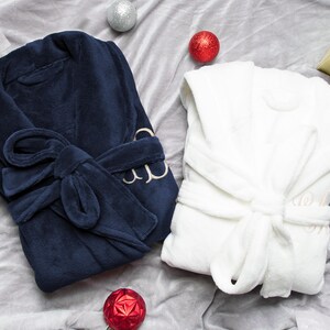 May include: Two plush bathrobes, one navy blue and one white, with a gold embroidered monogram on each. The robes are laid out on a gray surface with red and gold ornaments.