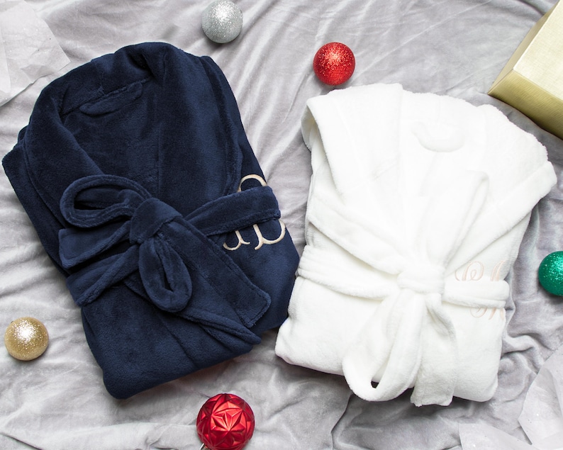 May include: Two plush bathrobes, one navy blue and one white, with a gold embroidered monogram on each. The robes are laid out on a gray surface with red and gold ornaments.
