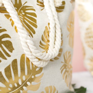 May include: A white canvas tote bag with gold foil monstera leaf print and white rope handles.