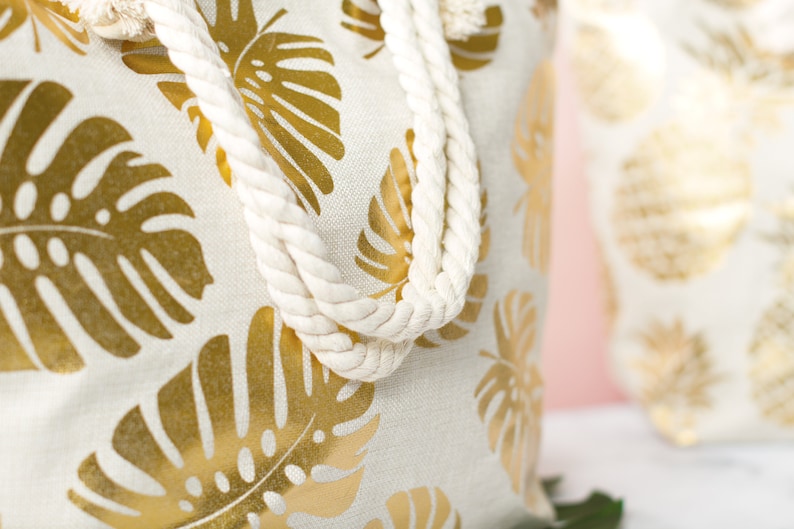 May include: A white canvas tote bag with gold foil monstera leaf print and white rope handles.