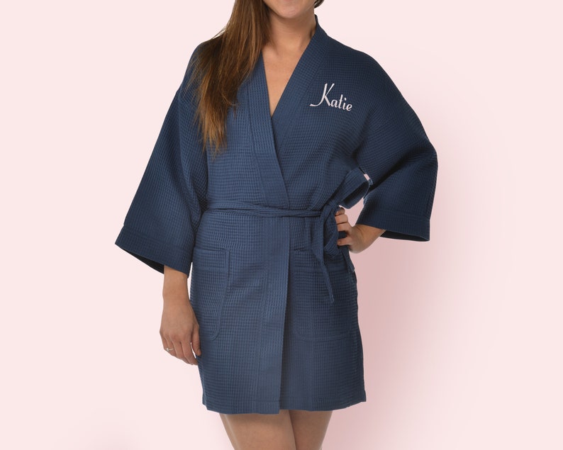 May include: A navy blue waffle-knit robe featuring a V-neckline, wide three-quarter length sleeves, and a matching tie belt at the waist. The front left chest area is embroidered with the name Katie in white cursive script lettering.