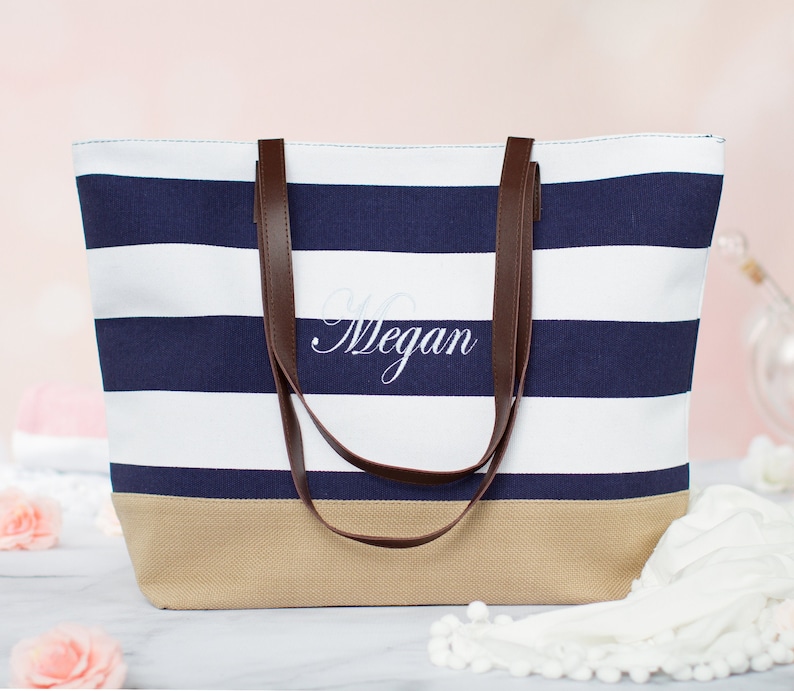 Puede incluir: Una bolsa de lona de rayas blancas y azul marino con asas de cuero marr&oacute;n. La bolsa tiene un fondo beige y el nombre "Megan" est&aacute; bordado en la parte delantera.