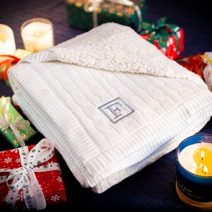 May include: A folded white knitted blanket with a cable knit design and a monogrammed letter "F" in a diamond shape. The blanket is surrounded by wrapped gifts and lit candles, suggesting a cozy, festive theme.
