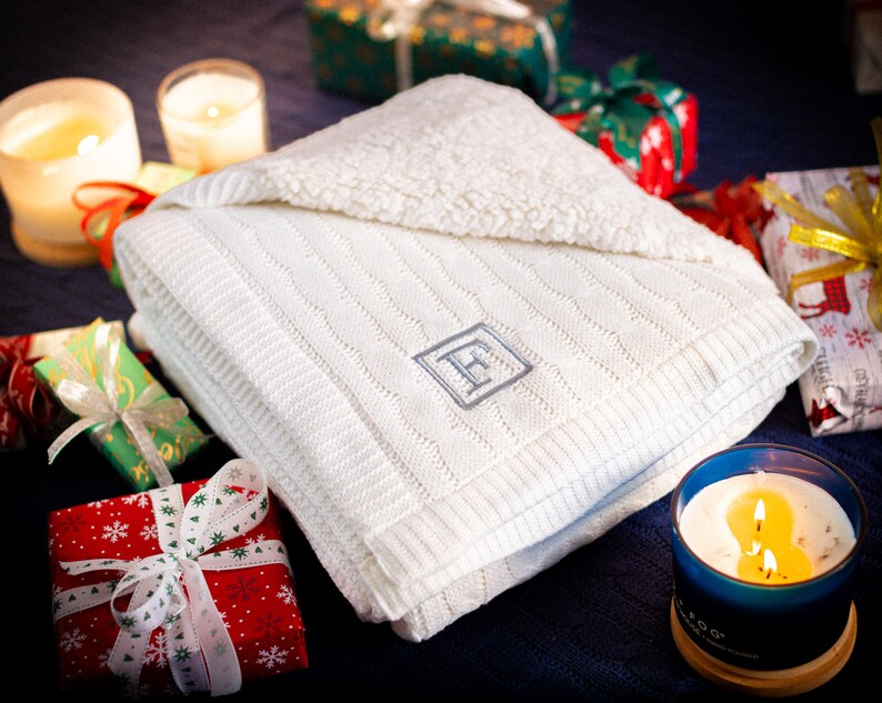 May include: A folded white knitted blanket with a cable knit design and a monogrammed letter "F" in a diamond shape. The blanket is surrounded by wrapped gifts and lit candles, suggesting a cozy, festive theme.