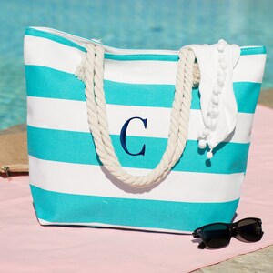 Puede incluir: Una bolsa de lona de rayas turquesa y blancas con asas de cuerda. La bolsa tiene una gran "C" bordada en la parte delantera. La bolsa est&aacute; sobre una superficie rosa con gafas de sol al lado.