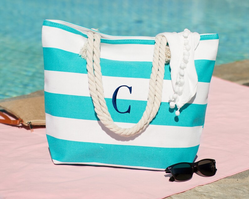Puede incluir: Una bolsa de lona de rayas turquesa y blancas con asas de cuerda. La bolsa tiene una gran "C" bordada en la parte delantera. La bolsa est&aacute; sobre una superficie rosa con gafas de sol al lado.