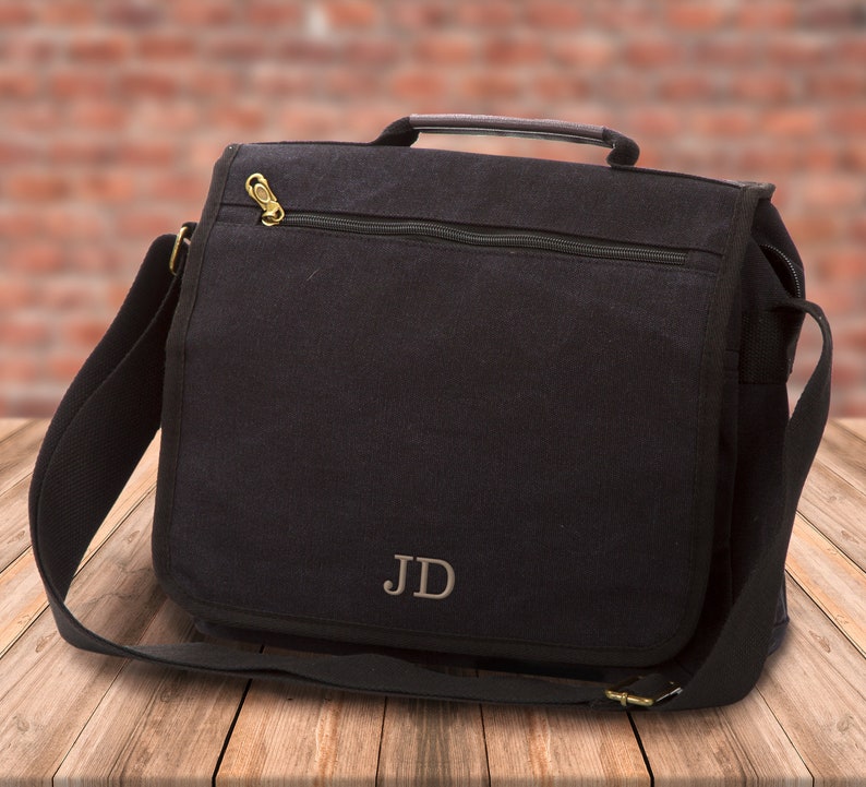 May include: A black canvas messenger bag with a zippered closure and a top handle. The bag has a personalized monogram "JD" on the front.