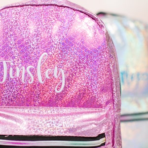 May include: Pink iridescent glitter backpack with a white embroidered name "Tinsley" on the front.