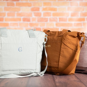 May include: Three nylon tote bags in light gray, rust, and pale pink, each with a shoulder strap. The light gray bag has a monogrammed "G". The rust bag has text. The bags are on a wooden surface.