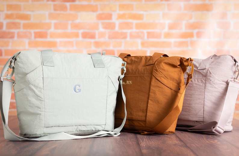 May include: Three nylon tote bags in light gray, rust, and pale pink, each with a shoulder strap. The light gray bag has a monogrammed "G". The rust bag has text. The bags are on a wooden surface.