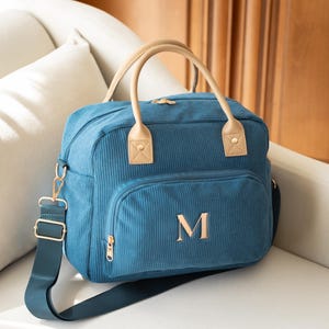 May include: Teal corduroy diaper bag with a front pocket and a gold zipper. The bag has tan leather handles and the letter "M" embroidered on the front. It also has a matching teal shoulder strap with gold hardware.