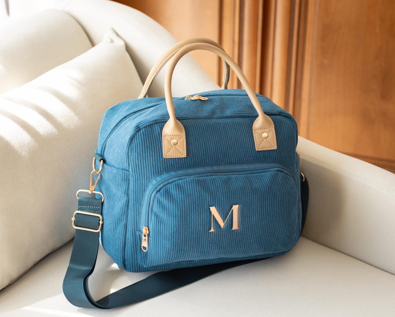 May include: Teal corduroy diaper bag with a front pocket and a gold zipper. The bag has tan leather handles and the letter "M" embroidered on the front. It also has a matching teal shoulder strap with gold hardware.