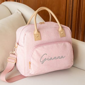 May include: A pink corduroy diaper bag with a top handle and a shoulder strap. The bag has a front pocket with the name "Gianna" embroidered in cursive. The bag has tan leather accents and gold-colored hardware.