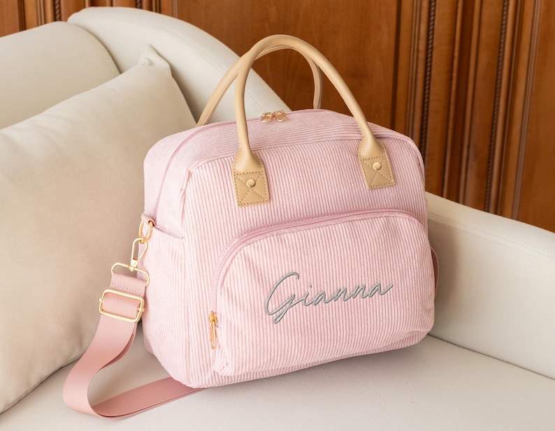 May include: A pink corduroy diaper bag with a top handle and a shoulder strap. The bag has a front pocket with the name "Gianna" embroidered in cursive. The bag has tan leather accents and gold-colored hardware.
