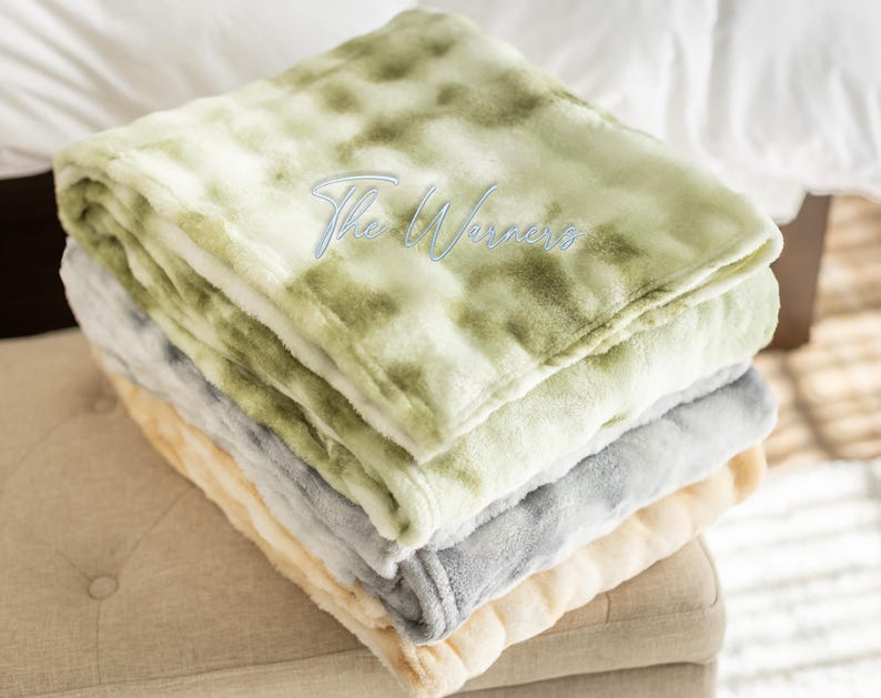 May include: A stack of four plush blankets in shades of green, gray, and cream. The top blanket has a tie-dye pattern. The blankets are folded neatly and stacked on top of each other. The text "The Warners" is embroidered on the top blanket.