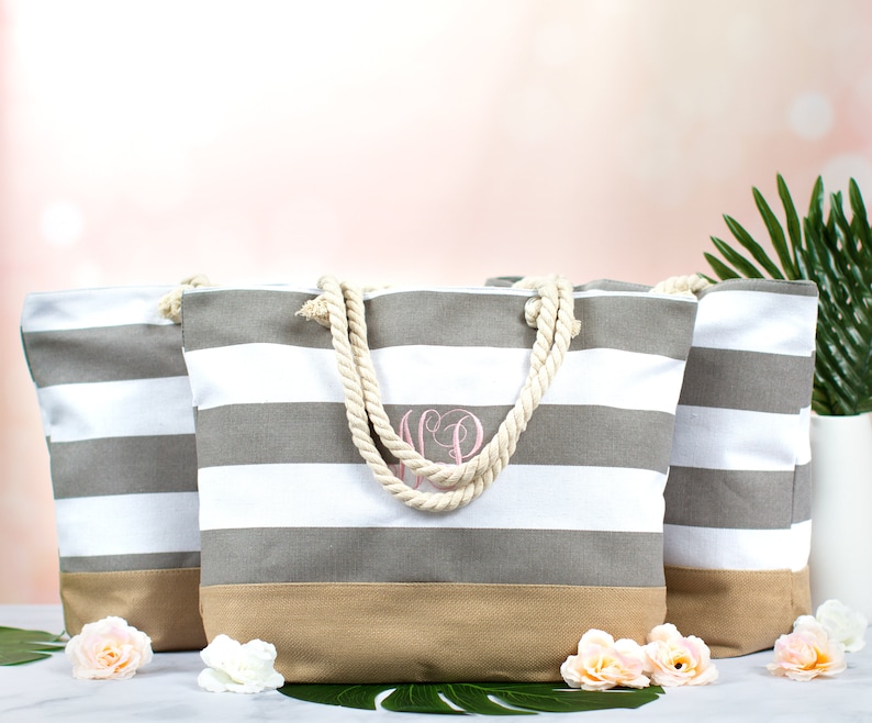 May include: Three striped tote bags with rope handles. The bags have gray and white horizontal stripes, a tan bottom, and a pink monogram. The bags are decorated with faux flowers and greenery.